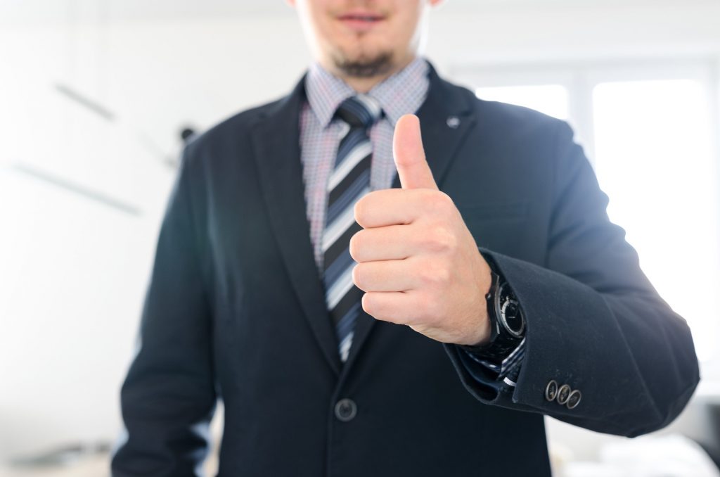 Homem de relógio, camisa, gravata listrada e terno azul fazendo o sinal de positivo com a mão esquerda, mostrando o employer branding positivo